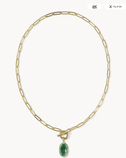 A gold paperclip chain necklace with a toggle clasp and a small oval green gemstone pendant, shown on a light gray background.