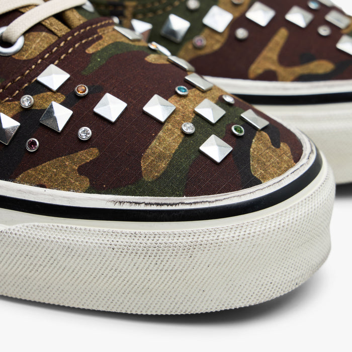 Vans LX Authentic 44. Close-up of silver metal pyramid studs on woodland camo canvas upper and distressed rubber foxing tape.
